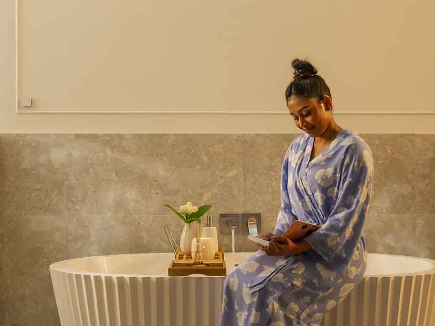 A woman seats on the side of an elegant white bathtub, reading a book as she waits for it to fill up