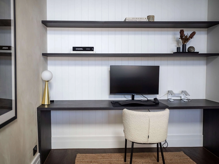 Modern home office design featuring a sleek black desk with a computer setup, stylish floating shelves with decorative objects, and a cozy chair set against a white paneled wall.