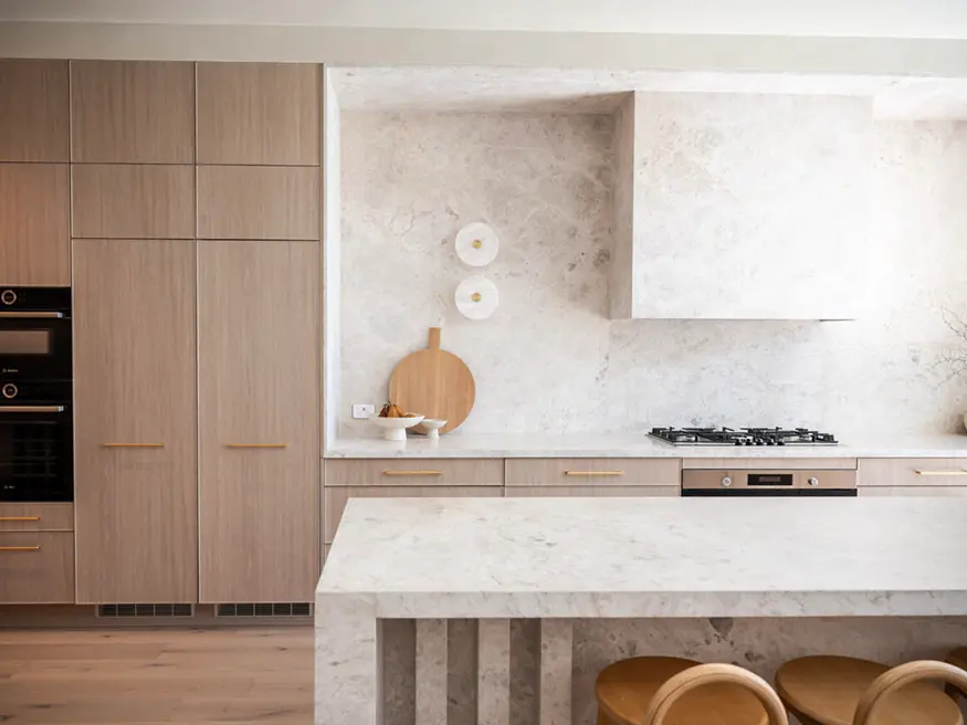 A minimalist kitchen with a stovetop, a wooden cutting board, and a metal faucet. Light enters through a window on the left.