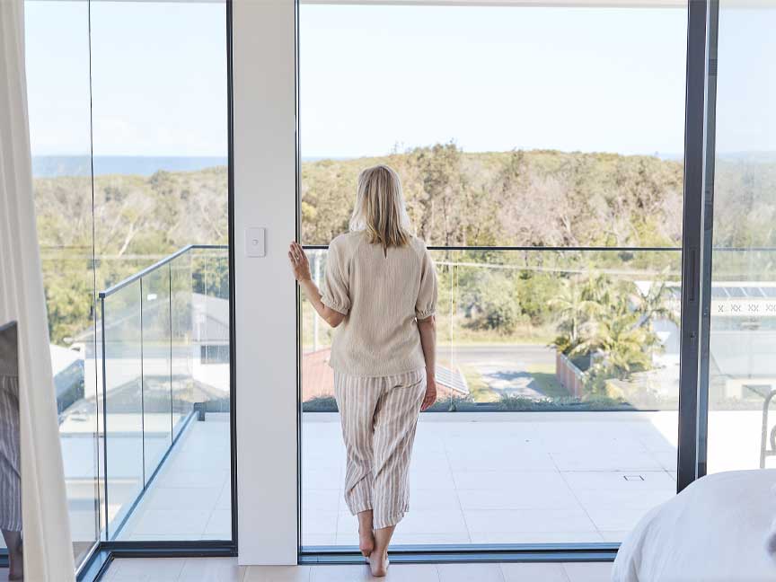 A woman stands on a balcony overlooking a scenic ocean view. She is wearing a linen top and striped pants and is looking out at the horizon.