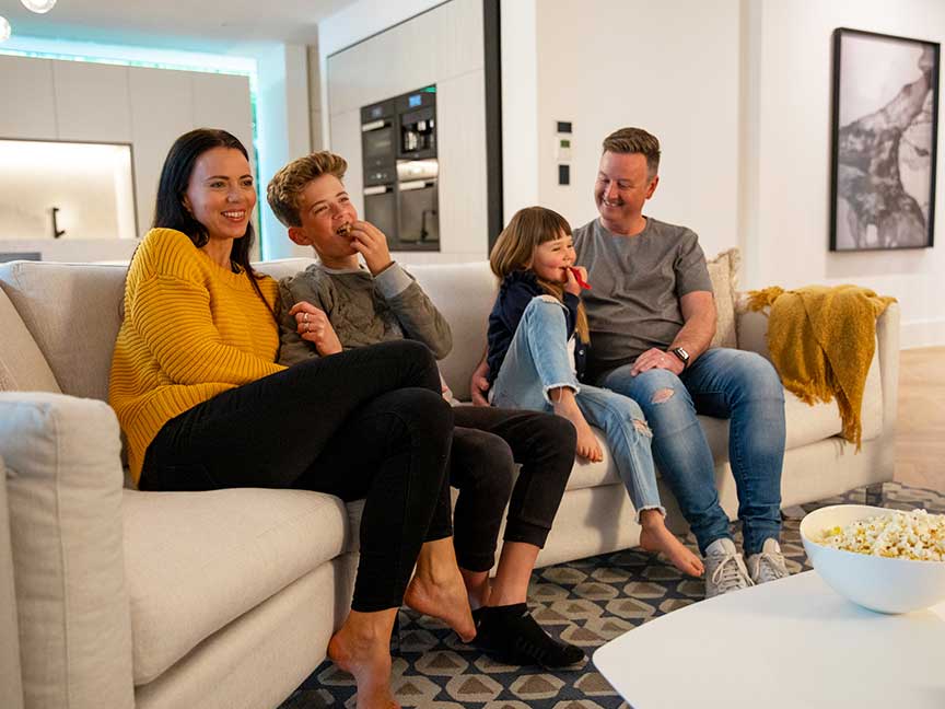 A smiling family sits together on a sofa at home, watching a funny movie on TV